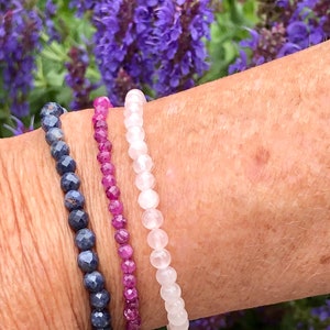 May include: Three beaded bracelets stacked on a wrist. The top bracelet is white, the middle bracelet is pink, and the bottom bracelet is blue.