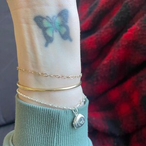 Peut inclure: Un poignet avec un tatouage de papillon et trois bracelets. Le bracelet du haut est une chaîne en or délicate, le bracelet du milieu est une bande épaisse en or et le bracelet du bas est une chaîne en argent avec un charme rond en argent.
