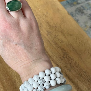 Puede incluir: Una mano con un anillo de plata con una piedra ovalada verde, y tres pulseras de cuentas blancas apiladas en la mu&ntilde;eca, con una pulsera de brazalete azul claro en la parte superior.