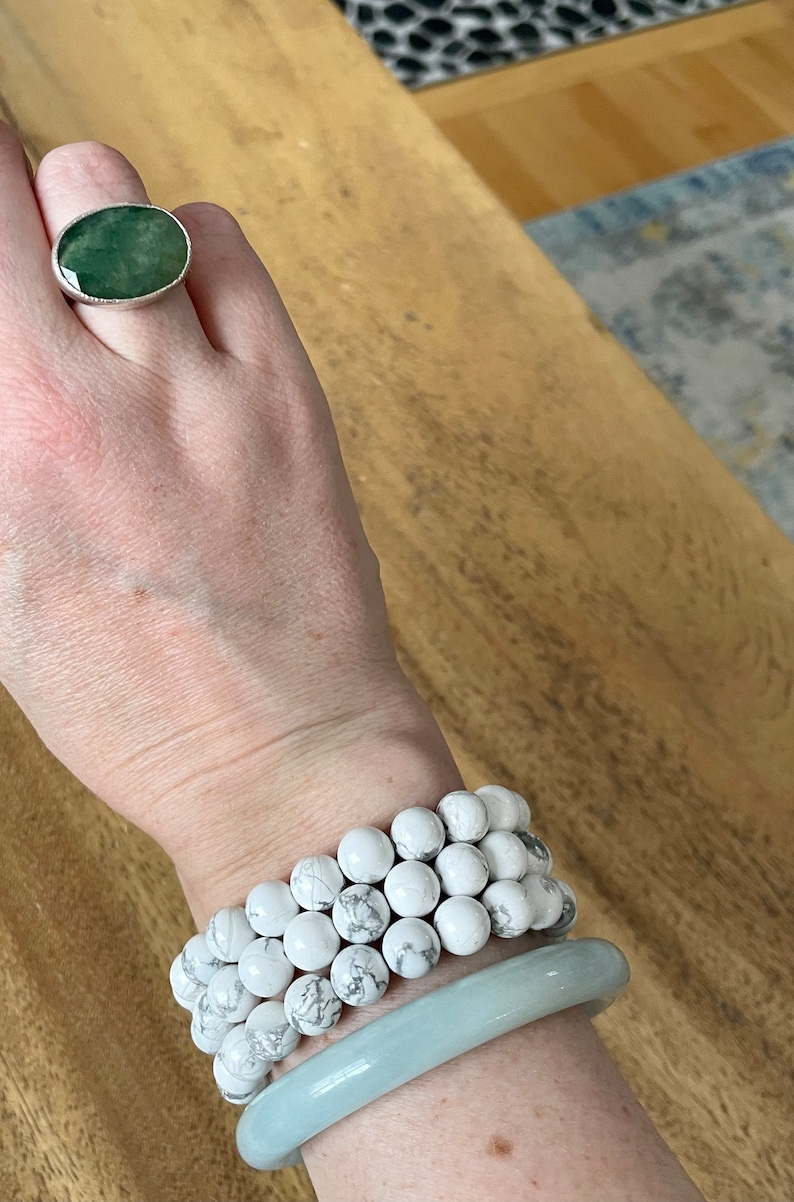Puede incluir: Una mano con un anillo de plata con una piedra ovalada verde, y tres pulseras de cuentas blancas apiladas en la mu&ntilde;eca, con una pulsera de brazalete azul claro en la parte superior.