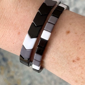 Peut inclure: Deux bracelets empilés en céramique noire, blanche et grise. Le bracelet du haut a un motif en chevron, tandis que le bracelet du bas a un motif carré.