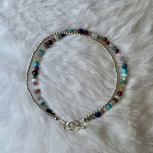 May include: A silver chain bracelet with a toggle clasp and colourful gemstone beads. The beads are in shades of pink, blue, green, purple, and brown.