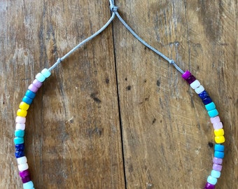 May include: A colorful beaded necklace with a gray cord. The beads are square and come in shades of yellow, blue, pink, purple, and white.