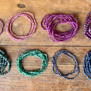 May include: Eight strands of gemstone beads are shown in an overhead view. The colours include pink, purple, green, and blue. The beads are arranged in circular patterns on a wooden surface.