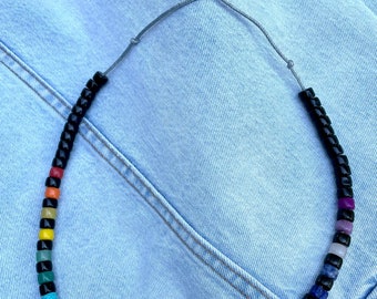 May include: A rainbow colored beaded necklace with black beads interspersed throughout. The necklace is made of small, round beads and is strung on a gray cord.