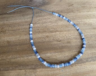 May include: A necklace made of blue and white glass beads. The beads are square shaped and have a mottled pattern. The necklace is tied with a gray cord.