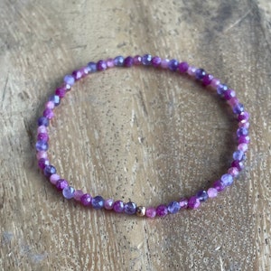 May include: A delicate bracelet made with small pink, purple and blue gemstone beads. A single gold bead is placed in the centre of the bracelet.