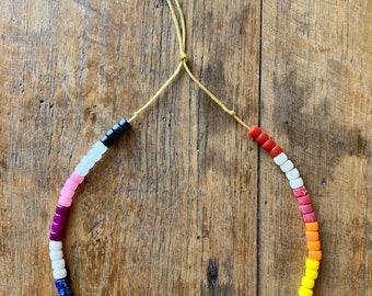 May include: A colorful beaded necklace with a rainbow of colors, including pink, red, orange, yellow, green, blue, and purple. The necklace is made of small, square beads and is strung on a thin, yellow cord.