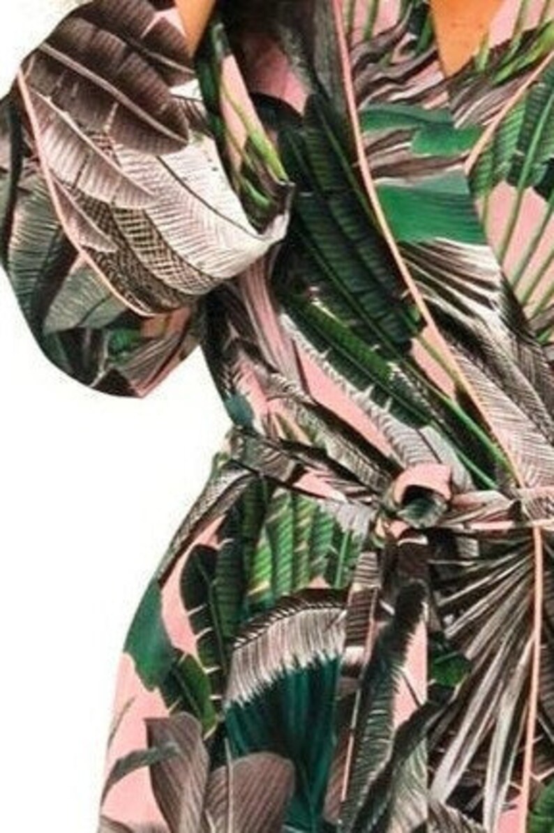 Puede incluir: Una bata de color rosa y verde con estampado tropical y una cinta para atar. La bata tiene manga larga y cuello en V.