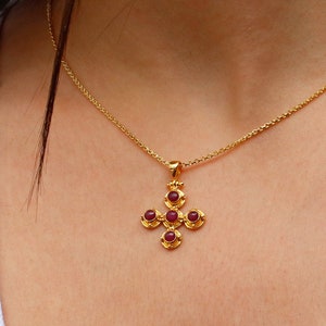 May include: A gold necklace with a small cross pendant featuring five red gemstones. The cross is ornate and has a textured design.