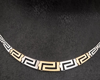 May include: A two-tone necklace featuring a repeating Greek key pattern. The chain consists of alternating silver-colored and gold-colored metal links, with the central section showcasing a prominent gold-colored Greek key motif. The brand name DIMOS is visible in the bottom right corner.