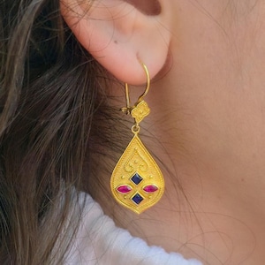 May include: Gold-toned teardrop earring with intricate filigree design, featuring red and blue gemstone accents. The earring is suspended from a hook and is shown against a blurred background. The earring has a decorative top.