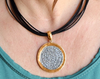 Pode incluir: Um colar com um pingente redondo de prata e ouro, com um design circular que apresenta letras intrincadas. O colar é usado em um cordão preto.