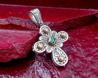 May include: A silver and gold cross pendant with a green gemstone and clear stones. The pendant is on a red background.