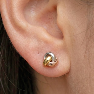 Può includere: Primo piano di un orecchino a nodo in oro, argento e oro rosa. L'orecchino è composto da tre anelli ad incastro, con finitura lucida. L'orecchino è indossato su un orecchio. Il marchio "DIMOS" si trova nell'angolo in basso a destra.