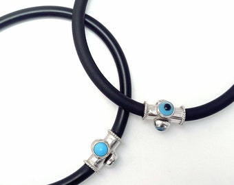 May include: Two black rubber bracelets with silver charms featuring blue evil eye beads. The charms are in the shape of a cylinder with a small bead on each side. The bracelets are on a white background.