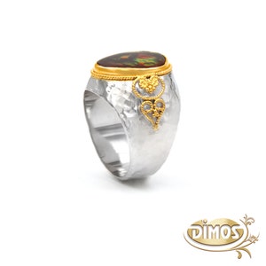 May include: A silver ring with a hammered finish and a gold band. The ring features a large oval-shaped gemstone with a brown and green pattern. The gold band is decorated with a delicate filigree design.