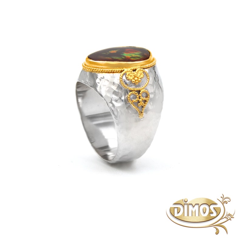 May include: A silver ring with a hammered finish and a gold band. The ring features a large oval-shaped gemstone with a brown and green pattern. The gold band is decorated with a delicate filigree design.