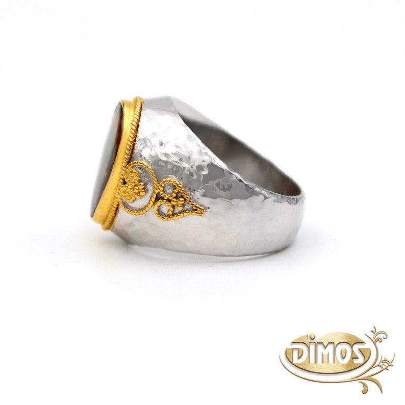 May include: A silver ring with a hammered finish and an oval-shaped gemstone set in a gold bezel. The gemstone has a red, brown, and white pattern. The ring has a gold filigree design on one side.
