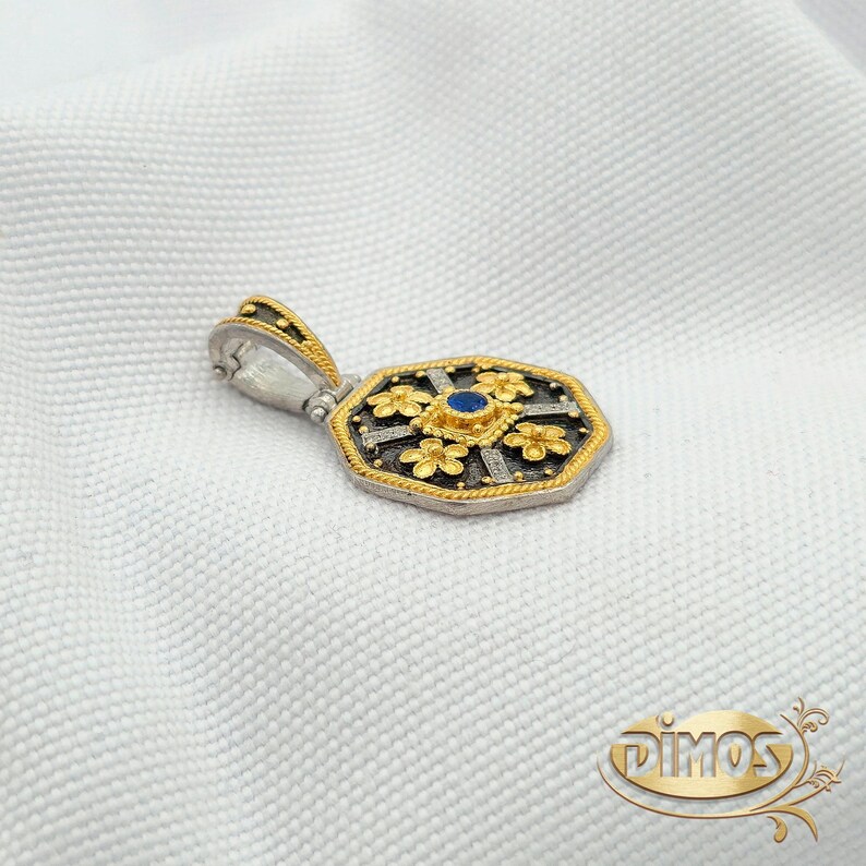 May include: A silver and gold pendant with an octagonal shape, featuring a central blue stone and floral accents. The pendant has a gold bail and is set against a white background.