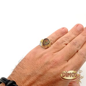 May include: A silver ring with a gold oval-shaped stone set in the centre. The stone has a floral design in brown, yellow, and red colours. The ring is on a person's finger.