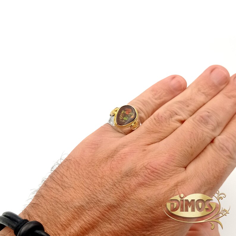 May include: A silver ring with a gold oval-shaped stone set in the centre. The stone has a floral design in brown, yellow, and red colours. The ring is on a person's finger.