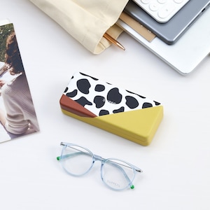 May include: A rectangular eyeglasses case with a cow-print design, brown accent, and yellow base sits near a pair of light blue eyeglasses. The glasses have a thin silver frame and the text "SUPPEAL" on the lens. A beige tote bag and a laptop are in the background.