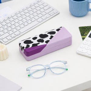 May include: A light purple eyeglasses case with a black and white cow print design, a purple accent, and a pair of light blue eyeglasses with green accents. A white keyboard and a blue mug are also visible.