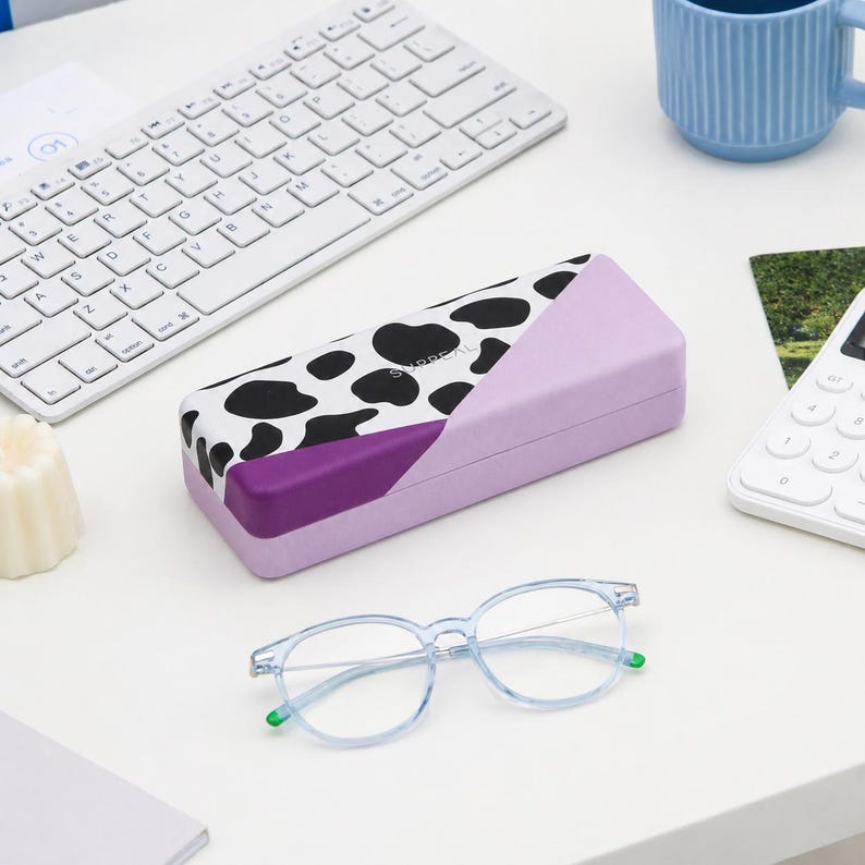 May include: A light purple eyeglasses case with a black and white cow print design, a purple accent, and a pair of light blue eyeglasses with green accents. A white keyboard and a blue mug are also visible.
