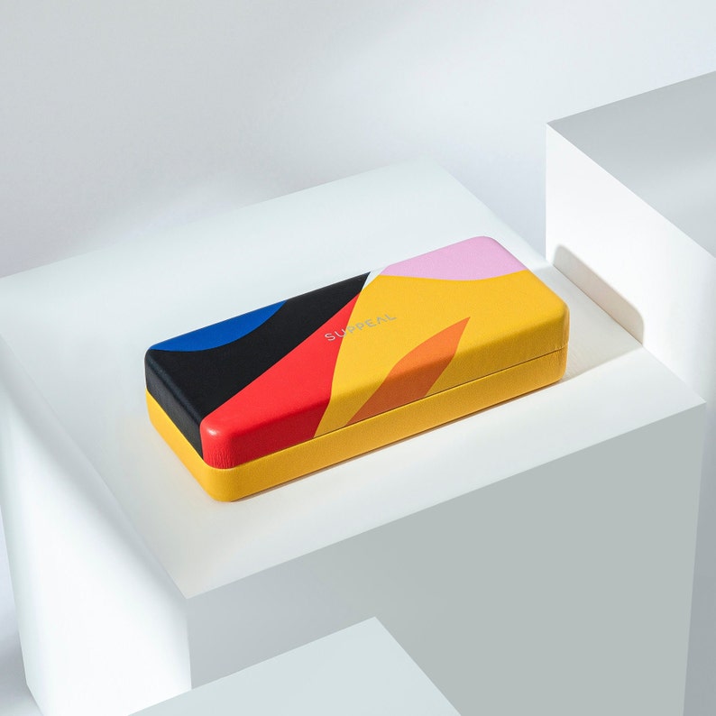 May include: A rectangular glasses case with a vibrant abstract design. The case features a yellow base and lid, with a colorful pattern of black, red, blue, pink, and orange shapes. The word "SURREAL" is printed in gold on the lid.