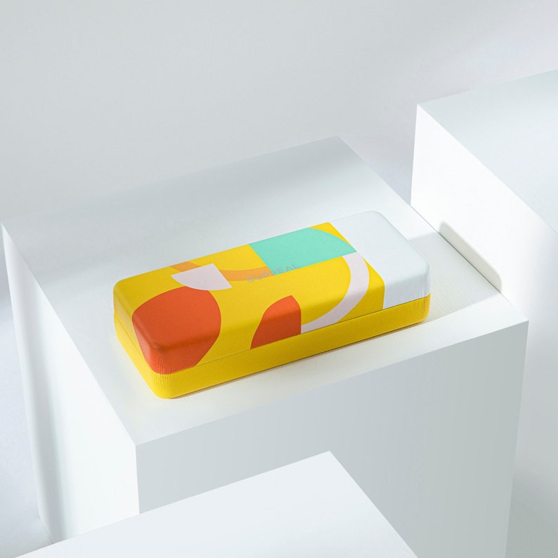 May include: A rectangular yellow case with a colorful abstract design. The design includes orange, teal, and white shapes. The case is resting on a white geometric surface.