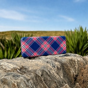 May include: A rectangular blue case with a plaid pattern in pink, white, and navy blue. The case is resting on a textured rock, with green plants and a blue sky in the background.
