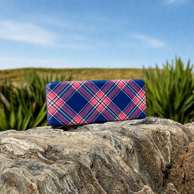 May include: A rectangular blue case with a plaid pattern in pink, white, and navy blue. The case is resting on a textured rock, with green plants and a blue sky in the background.