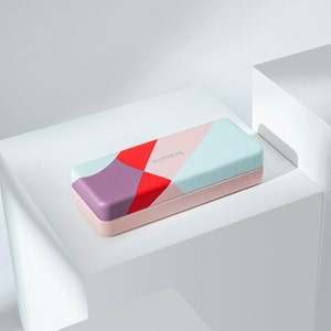May include: A rectangular case with a geometric design in red, purple, and blue-green. The case has a light pink base and the word "SUPPEAL" is printed on the top. The case is resting on a white surface.