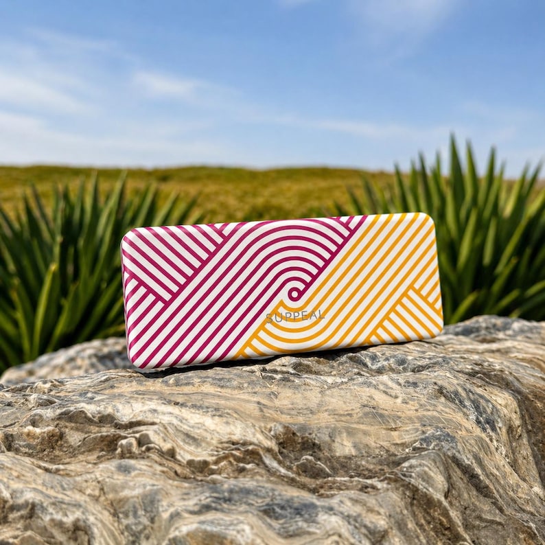 May include: Rectangular case with a geometric pattern of white, burgundy, and yellow lines. The word "SUPPEAL" is printed on the case. The case is resting on a rock with a natural background of green plants and a blue sky.