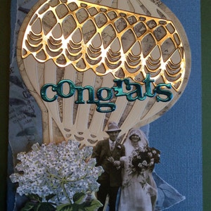 May include: A card with a gold and white hot air balloon design with the word "congrats" in teal lettering. The card features a couple in vintage clothing, a bouquet of flowers, and a floral design.