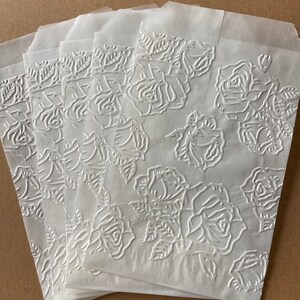 May include: A stack of six white paper bags with a raised embossed rose pattern. The bags are folded and stacked on a brown surface.
