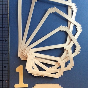 May include: A set of eight white paper frames with a stitched border, arranged in a fan shape. The frames are layered on top of each other, with the largest frame at the bottom and the smallest frame at the top. A single white paper frame with a stitched border is below the fan of frames. A white paper number one is to the left of the frames.