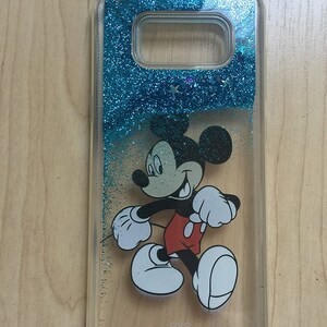 May include: A clear phone case with a blue glitter background and a Mickey Mouse character walking on the front. The case has a clear back and a blue glitter top. The case has a small hole for the camera lens.