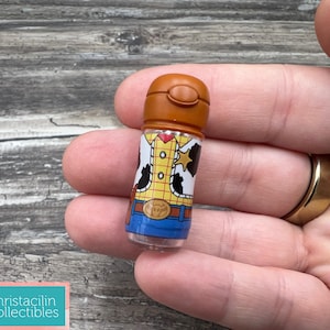 May include: A small, cylindrical collectible featuring a design inspired by the character Woody from the movie Toy Story. The item has a brown cap and a clear body, with Woody's outfit printed on it. The collectible is held in a hand.