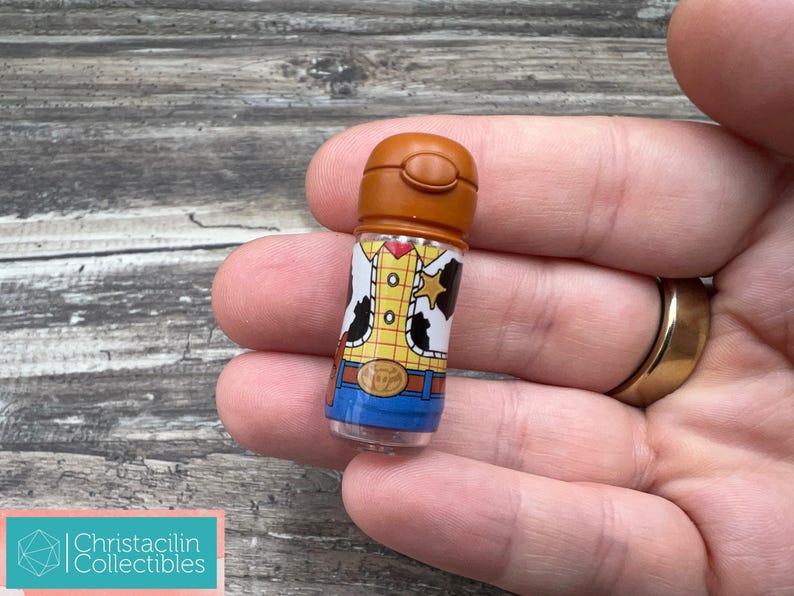 May include: A small, cylindrical collectible featuring a design inspired by the character Woody from the movie Toy Story. The item has a brown cap and a clear body, with Woody's outfit printed on it. The collectible is held in a hand.