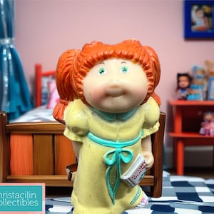 May include: A vintage Cabbage Patch Kids doll with red hair in pigtails, wearing a yellow dress with a blue sash, holding a small book. The doll is in a child's bedroom setting with a small bed and other toys.