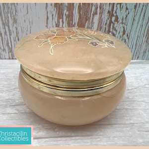 May include: A peach-colored, round trinket box with a floral design on the lid. The box has a gold-colored metal band around the middle. The lid is slightly domed and the base is rounded. The box is made of a smooth, glossy material.