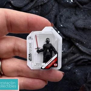 May include: A Kylo Ren action figure from Star Wars in a clear plastic package. The figure is black and holds a red lightsaber. The package has the Star Wars Toybox logo and the number 13.