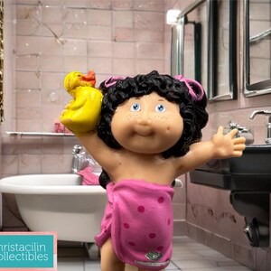 May include: A Cabbage Patch Kids doll figurine in a bathroom setting. The doll has black curly hair, blue eyes, and is wrapped in a pink towel with polka dots. It holds a yellow rubber duck. The background includes a sink, tub, and mirror.
