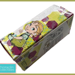 May include: A rectangular box featuring a cartoon image of a young girl with blonde hair and a green dress. The box has a yellow, maroon, and white geometric pattern. The box is labeled "Christacilin Collectibles".