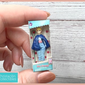 May include: A miniature Bo Peep doll in a box. The doll is wearing a blue cape, a pink sash, and blue pants with ruffled trim. The box has a light blue top with a flower and the words "Bo Peep" on the front.