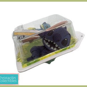 May include: A blue Stitch figure in a clear plastic package. The figure has a large grin and is lying on a green surface. The package has the word "Stitch" and the number "13" printed on it. The figure is a collectible.