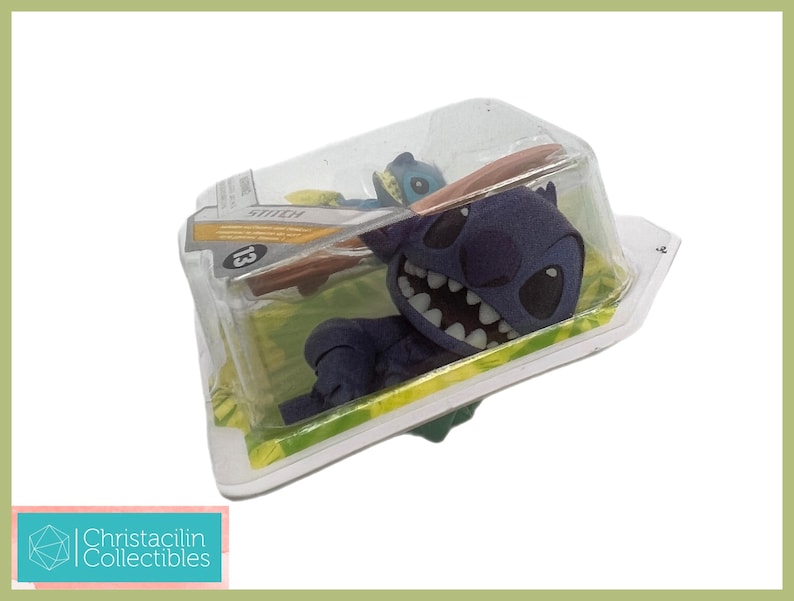 May include: A blue Stitch figure in a clear plastic package. The figure has a large grin and is lying on a green surface. The package has the word "Stitch" and the number "13" printed on it. The figure is a collectible.