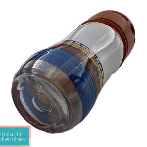 May include: A clear glass bottle with a brown cap. The bottle has a patterned design with blue, white, and yellow stripes. The bottle is cylindrical and appears to be a decorative item.